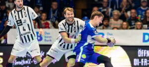 Zwei Handballspieler in schwarz-weißen Trikots verteidigen sich gegen einen Spieler in einem blauen Trikot, der den Ball hält, während im Hintergrund eine Zuschauermenge zusieht.