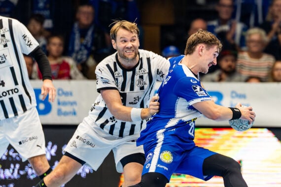 Zwei Handballspieler in schwarz-weißen Trikots verteidigen sich gegen einen Spieler in einem blauen Trikot, der den Ball hält, während im Hintergrund eine Zuschauermenge zusieht.