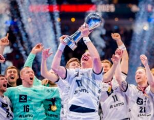 Eine jubelnde Handballmannschaft in weißen Trikots feiert, während ein Spieler eine große Trophäe über seinen Kopf hebt, umgeben von jubelnden Mitspielern und Konfetti in einer Arena.