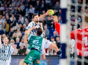 Ein Handballspieler in einem weiß-schwarz gestreiften Trikot springt zum Schuss, während ein Verteidiger in einem grünen Trikot den Schuss abblockt, während im Hintergrund ein Torwart in Rot und eine verschwommene Menschenmenge zu sehen sind.