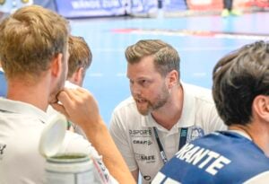 Ein Trainer in einem weißen Poloshirt spricht während eines Handballspiels intensiv mit seiner Mannschaft, während die Spieler auf der Bank aufmerksam zuhören. Die Szene findet in einer Halle auf einem blauen Spielfeld statt.