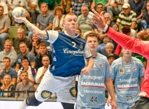 Ein männlicher Handballspieler in einem blauen Trikot springt, um den Ball zu werfen, während Verteidiger und ein Torwart in Rot und Blau versuchen, ihn zu blockieren. Eine Menschenmenge beobachtet das intensive Spiel im Hintergrund.