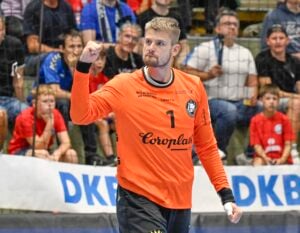 Ein männlicher Handballtorwart in einem orangefarbenen Trikot mit der Nummer 1 hebt jubelnd die Faust. Die Zuschauer auf der Tribüne verfolgen das Geschehen, und im Hintergrund ist ein DKB-Banner zu sehen.