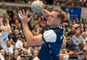 Ein männlicher Handballspieler in einem blauen Trikot bereitet sich darauf vor, den Ball während eines Spiels zu werfen, während im Hintergrund eine konzentrierte Menge zuschaut.