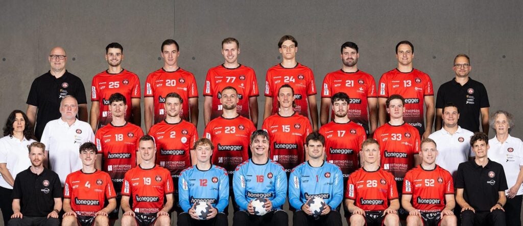 25_Vor_BSVBern_Team Eine Männerhandballmannschaft in roten Trikots posiert für ein Gruppenfoto mit Trainern und Mitarbeitern in schwarzen oder weißen Hemden, die in drei Reihen vor einem schlichten grauen Hintergrund stehen und sitzen. Zwei Torhüter in Blau halten Handbälle in der Hand.