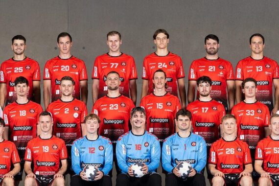 Eine Männerhandballmannschaft in roten Trikots posiert für ein Gruppenfoto mit Trainern und Mitarbeitern in schwarzen oder weißen Hemden, die in drei Reihen vor einem schlichten grauen Hintergrund stehen und sitzen. Zwei Torhüter in Blau halten Handbälle in der Hand.