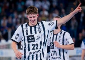 Ein lächelnder männlicher Handballspieler in einem schwarz-weiß gestreiften Trikot mit der Nummer 22 jubelt mit erhobenem Arm. Ein anderer Mitspieler ist teilweise hinter ihm auf dem Spielfeld zu sehen.