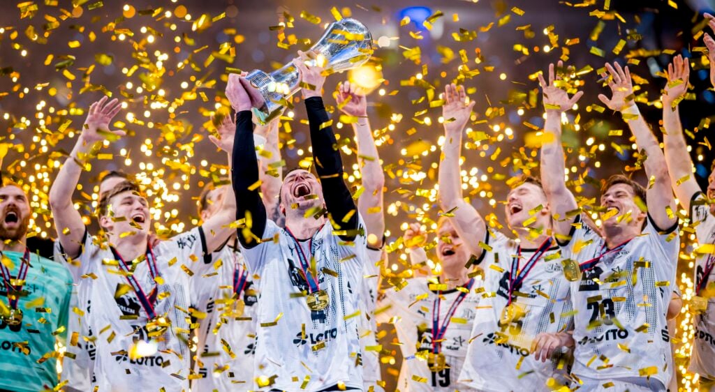 Eine jubelnde Sportmannschaft in weißen Trikots feiert einen Meisterschaftssieg und hebt eine Trophäe inmitten von fallendem goldenem Konfetti in die Höhe. Die Spieler jubeln mit erhobenen Armen und tragen Medaillen um ihren Hals.