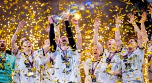 Eine jubelnde Sportmannschaft in weißen Trikots feiert einen Meisterschaftssieg und hebt eine Trophäe inmitten von fallendem goldenem Konfetti in die Höhe. Die Spieler jubeln mit erhobenen Armen und tragen Medaillen um ihren Hals.