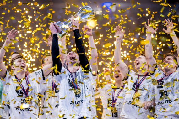 Eine jubelnde Sportmannschaft in weißen Trikots feiert einen Meisterschaftssieg und hebt eine Trophäe inmitten von fallendem goldenem Konfetti in die Höhe. Die Spieler jubeln mit erhobenen Armen und tragen Medaillen um ihren Hals.