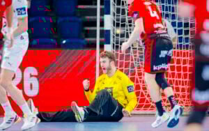 Ein Handballtorwart in einem gelben Trikot sitzt auf dem Boden und jubelt, während sich zwei gegnerische Spieler in roten und schwarzen Trikots in der Nähe auf dem Spielfeld bewegen. Im Hintergrund sind ein Tornetz und eine Anzeigetafel zu sehen.