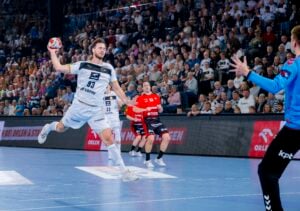 Ein Handballspieler in einem weißen Trikot springt, um den Ball in Richtung Tor zu werfen, während ein Torwart in Blau sich darauf vorbereitet, den Ball zu blocken. Zwei Gegner in Rot und Schwarz schauen zu. Die Arena ist mit Zuschauern gefüllt.