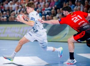Ein Handballspieler in einem weißen Trikot rennt mit dem Ball und weicht einem Verteidiger in einem roten Trikot aus, der versucht, ihn aufzuhalten. Die Aktion findet auf einem Hallenplatz statt, mit einer unscharfen Menschenmenge im Hintergrund.