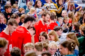 Eine Gruppe von Sportlern in roten Hemden steht inmitten einer großen Menge aufgeregter Kinder und Fans, von denen viele Gegenstände für Autogramme in einer lebhaften Halle bereithalten.