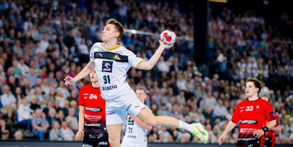 Ein Handballspieler in einem weißen Trikot springt in die Luft, um den Ball zu werfen, während zwei gegnerische Spieler in roten Trikots während eines Spiels in einer überfüllten Arena zusehen.