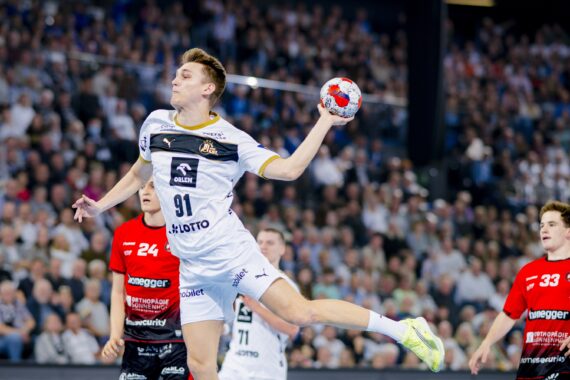 Ein Handballspieler in einem weißen Trikot springt in die Luft, um den Ball zu werfen, während zwei gegnerische Spieler in roten Trikots während eines Spiels in einer überfüllten Arena zusehen.