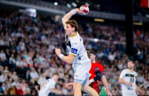 Ein Handballspieler in einem weißen Trikot bereitet sich während eines Spiels auf den Wurf vor, während im Hintergrund Zuschauer und gegnerische Spieler in roten Trikots zu sehen sind.