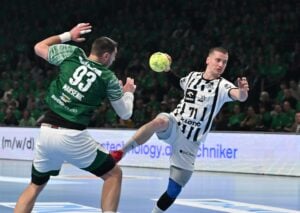 Ein Handballspieler in einem schwarz-weißen Trikot springt, um den Ball zu werfen, während ein Verteidiger in einem grünen Trikot versucht, ihn während eines Hallenspiels zu blockieren. Im Hintergrund sind Zuschauer zu sehen.