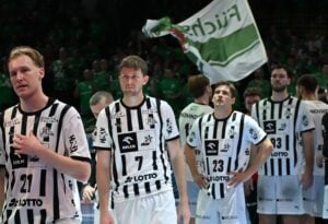 Eine Herren-Handballmannschaft in schwarz-weiß gestreiften Trikots steht auf einem Spielfeld und schaut ernst. Im Hintergrund schwenken Menschen in grünen Hemden eine grün-weiße Flagge. Die Atmosphäre wirkt angespannt oder feierlich.