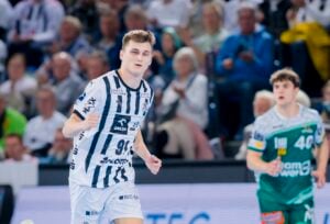 Ein männlicher Handballspieler in einem weiß-schwarz gestreiften Trikot läuft während eines Spiels auf einem Hallenplatz. Im unscharfen Hintergrund sind Zuschauer auf der Tribüne und ein weiterer Spieler in einem grünen Trikot zu sehen.