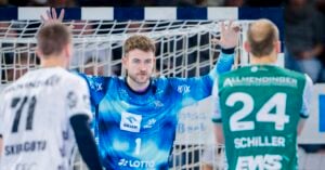 Ein Handballtorwart in einem blauen Trikot steht während eines Spiels mit erhobenen Armen aufmerksam vor zwei gegnerischen Spielern in weißen und grünen Trikots in der Nähe des Tornetzes.