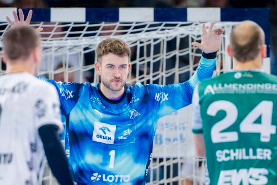 Ein Handballtorwart in einem blauen Trikot steht während eines Spiels mit erhobenen Armen aufmerksam vor zwei gegnerischen Spielern in weißen und grünen Trikots in der Nähe des Tornetzes.