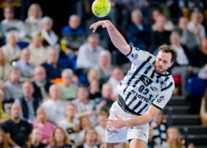 Ein männlicher Handballspieler in einem schwarz-weiß gestreiften Trikot springt und wirft einen gelben Ball während eines Spiels, während im Hintergrund ein unscharfes Publikum zusieht.