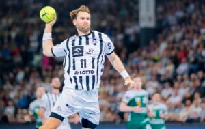 Ein Handballspieler in einem weiß-blau gestreiften Trikot springt in der Luft und bereitet sich darauf vor, einen gelben Ball zu werfen. Im Hintergrund sind eine verschwommene Menschenmenge und andere Spieler in grünen Trikots zu sehen.