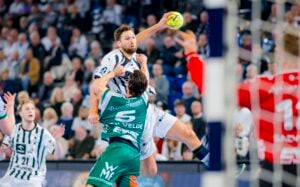 Ein Handballspieler in einem weiß-schwarzen Trikot springt, um den Ball zu werfen, während ein Verteidiger in einem grünen Trikot und ein Torwart in Rot versuchen, ihn während eines intensiven Spiels in einer überfüllten Halle zu blockieren.