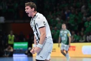 Ein männlicher Handballspieler in einem weiß-schwarzen Trikot ballt die Fäuste und schreit leidenschaftlich, während im Hintergrund unscharfe Zuschauer und ein weiterer Spieler zu sehen sind.
