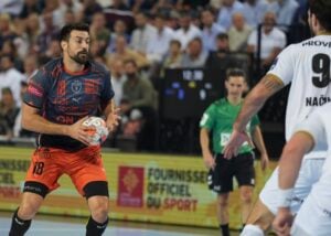 Ein Handballspieler in einem dunkelblauen und orangefarbenen Trikot hält den Ball und bereitet sich auf einen Pass oder einen Schuss vor, während er während eines Spiels einem Gegner in einem weißen Trikot gegenübersteht. Im Hintergrund sind Zuschauer und ein Schiedsrichter zu sehen.