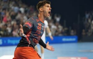 Ein männlicher Handballspieler in einem schwarz-orangenen Trikot feiert leidenschaftlich auf dem Spielfeld mit offenem Mund und weit ausgebreiteten Armen, während im Hintergrund eine verschwommene Menschenmenge zuschaut.