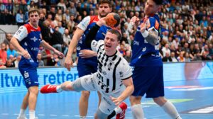 Ein männlicher Handballspieler in einem weißen Trikot springt nach vorne und hält den Ball, umgeben von gegnerischen Spielern in blauen Trikots, während die Fans auf der Tribüne einer überfüllten Halle zusehen.