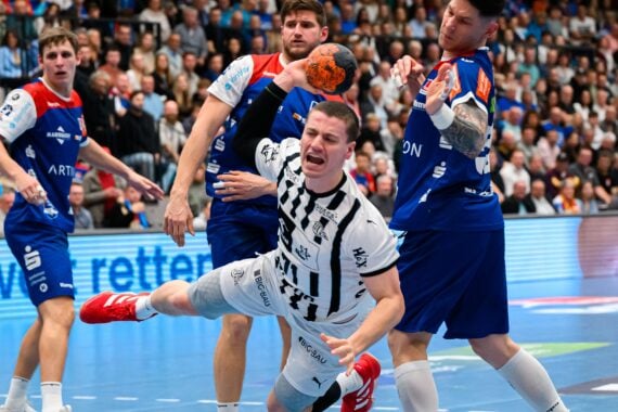 Ein männlicher Handballspieler in einem weißen Trikot springt nach vorne und hält den Ball, umgeben von gegnerischen Spielern in blauen Trikots, während die Fans auf der Tribüne einer überfüllten Halle zusehen.