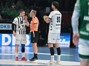 Ein Schiedsrichter spricht mit zwei Handballspielern in weißen und schwarzen Trikots auf einem Spielfeld, während ein weiterer Spieler in einem grünen Trikot in der Nähe steht, teilweise außerhalb des Bildes.