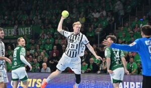Ein Handballspieler in einem schwarz-weiß gestreiften Trikot springt in der Luft, um den Ball zu werfen, umgeben von Spielern in grünen Trikots, mit einem dicht gedrängten Publikum im Hintergrund.