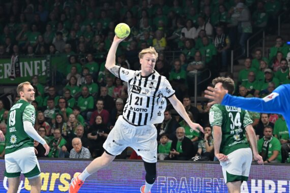Ein Handballspieler in einem schwarz-weiß gestreiften Trikot springt in der Luft, um den Ball zu werfen, umgeben von Spielern in grünen Trikots, mit einem dicht gedrängten Publikum im Hintergrund.
