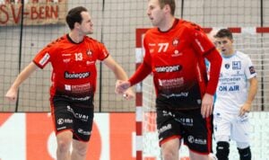 Zwei männliche Handballspieler in roten Trikots feiern auf dem Spielfeld, während ein Spieler in einem weißen Trikot im Hintergrund zusieht. Hinter ihnen sind ein Netz und Werbebanner zu sehen.