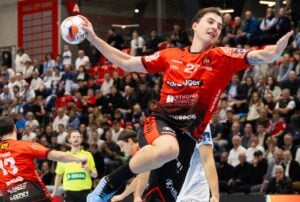 Ein Handballspieler in einem roten Trikot springt in der Luft und bereitet sich darauf vor, den Ball während eines Spiels zu werfen. Die Zuschauer schauen von der Tribüne im Hintergrund zu, und die gegnerischen Spieler in Weiß versuchen, ihn zu blockieren.