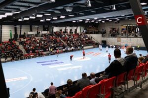 Eine große Halle mit Zuschauern, die ein Handballspiel verfolgen; die Spieler in roten und schwarzen Trikots stehen auf dem Spielfeld, und die Zuschauer füllen die Tribünen auf beiden Seiten des Spielfelds.