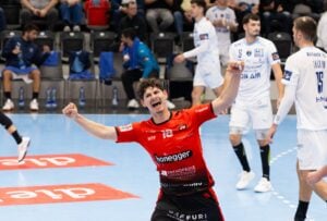 Ein männlicher Handballspieler in einem roten Trikot jubelt mit erhobenen Armen auf einem Hallenplatz, während im Hintergrund Spieler in weißen Trikots und Zuschauer zu sehen sind.