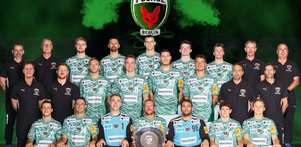 25_vor_Berlin_Team_Krickau Eine Männer-Handballmannschaft in grünen Camouflage-Trikots posiert mit Trainern und Betreuern in schwarzen Hemden und hält einen Pokalteller. Dahinter ist das Logo der Füchse Berlin auf grünem und schwarzem Hintergrund zu sehen - ein Hinweis auf die Ambitionen des THW Kiel in der Handball-Bundesliga Saison 2025/2026.