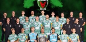 Eine Männer-Handballmannschaft in grünen Camouflage-Trikots posiert mit Trainern und Betreuern in schwarzen Hemden und hält einen Pokalteller. Dahinter ist das Logo der Füchse Berlin auf grünem und schwarzem Hintergrund zu sehen - ein Hinweis auf die Ambitionen des THW Kiel in der Handball-Bundesliga Saison 2025/2026.