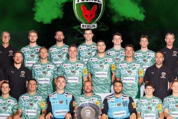 Eine Männer-Handballmannschaft in grünen Camouflage-Trikots posiert mit Trainern und Betreuern in schwarzen Hemden und hält einen Pokalteller. Dahinter ist das Logo der Füchse Berlin auf grünem und schwarzem Hintergrund zu sehen - ein Hinweis auf die Ambitionen des THW Kiel in der Handball-Bundesliga Saison 2025/2026.