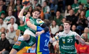 Ein Handballspieler in einem grün-weißen Trikot springt zum Schuss, wird aber von einem blauen Verteidiger geblockt. Ein anderer grüner Spieler schaut zu. Im Hintergrund ist eine jubelnde Menge zu sehen.