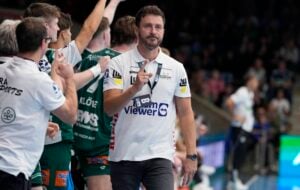 Ein Mann in einem weißen Polohemd mit Logos zeigt den Daumen nach oben, während er in einer Sporthalle zwischen Menschen in grünen Uniformen steht, wahrscheinlich Mannschaftskameraden oder Spieler. Im Hintergrund sind weitere Personen und unscharfe Zuschauerplätze zu sehen.