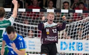 Ein Handballtorwart, der ein schwarzes Trikot mit der Nummer 12 trägt, jubelt energisch vor dem Tor und ballt beide Fäuste, während im Hintergrund andere Spieler und Zuschauer zu sehen sind.