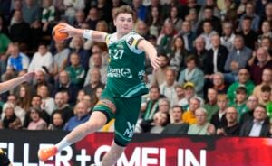 Ein männlicher Handballspieler in einem grünen Trikot springt in der Luft, hält einen orangefarbenen Ball und bereitet sich auf den Wurf vor. Eine große Menschenmenge im Hintergrund verfolgt das Geschehen mit Spannung.