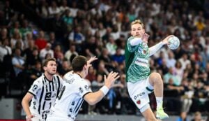 Ein Handballspieler in einem grün-weißen Trikot springt zum Wurf, als zwei Verteidiger des THW Kiel ihn herausfordern, während eines intensiven Spiels der Handball-Bundesliga Saison 2025/2026 gegen die Füchse Berlin, mit einer unscharfen Zuschauermenge im Hintergrund.