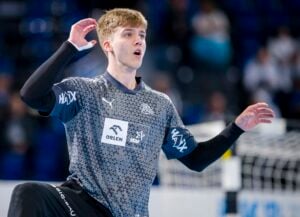 Ein junger männlicher Handballspieler in einem grauen Trikot hebt einen Arm und scheint während eines Spiels konzentriert zu sein. Im Hintergrund sind ein unscharfes Hallenfeld und ein Tor zu sehen.